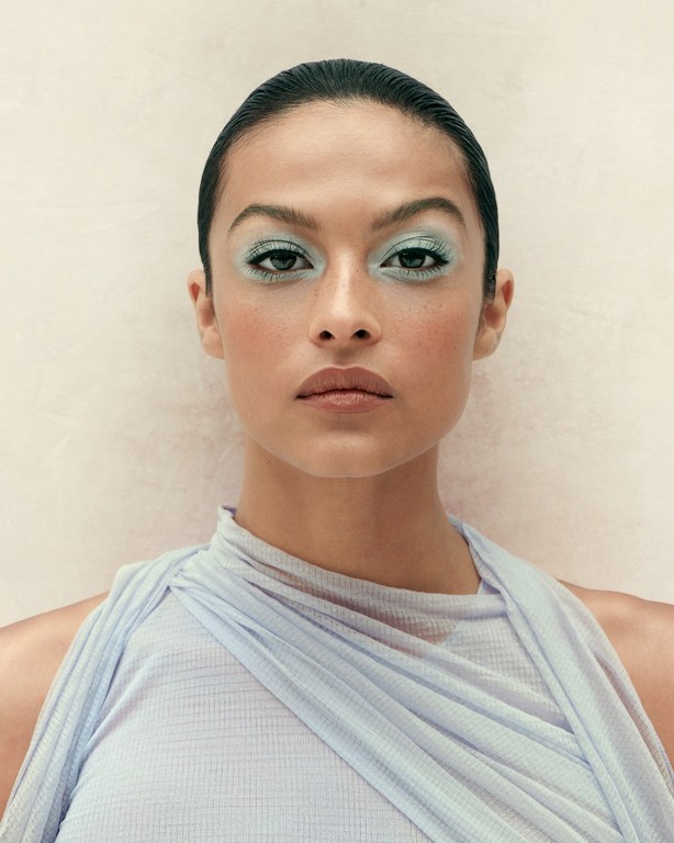 Close-up of a woman with light blue eyeshadow wearing a pale blue draped top against a light neutral background in a studio setting.