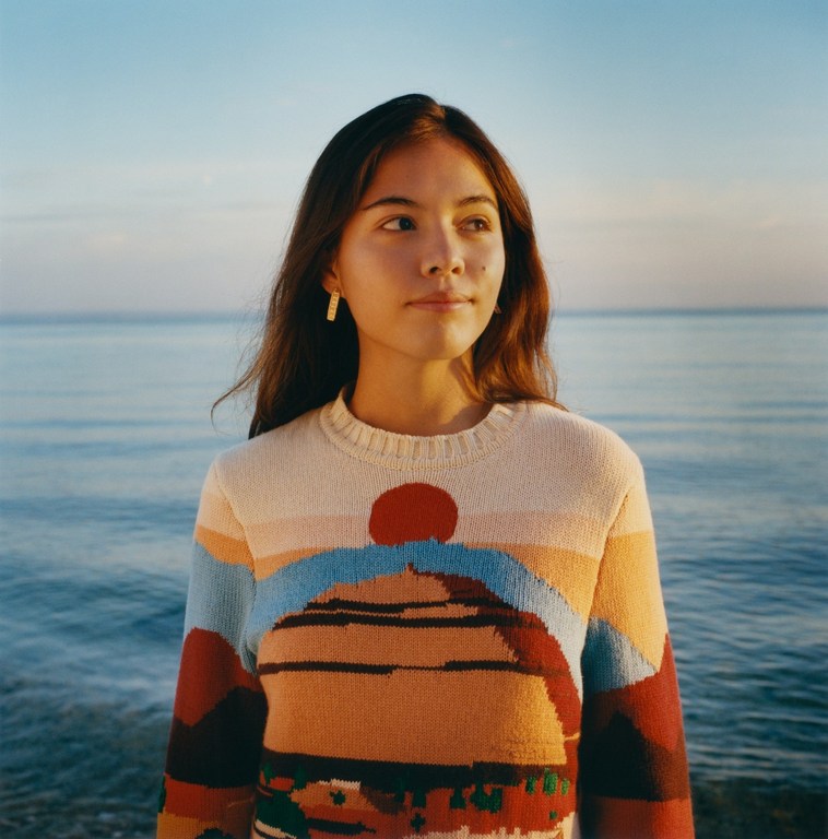 A young adult woman poses near the ocean at sunset wearing a colourful knit sweater with a landscape design and simple gold earrings.