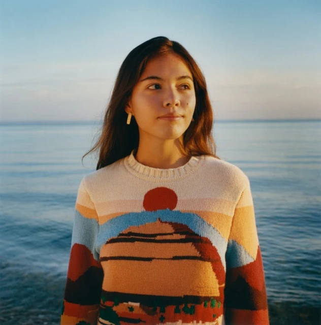A young adult woman poses near the ocean at sunset wearing a colourful knit sweater with a landscape design and simple gold earrings.