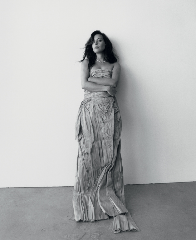 Monochrome portrait of a woman in a strapless draped gown leaning against a plain wall styled by Max Ortega
