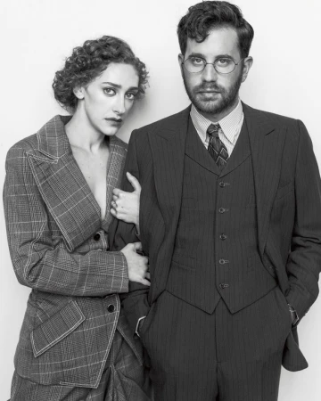 Black and white portrait of a woman in a plaid suit holding the arm of a man in a three piece pinstripe suit