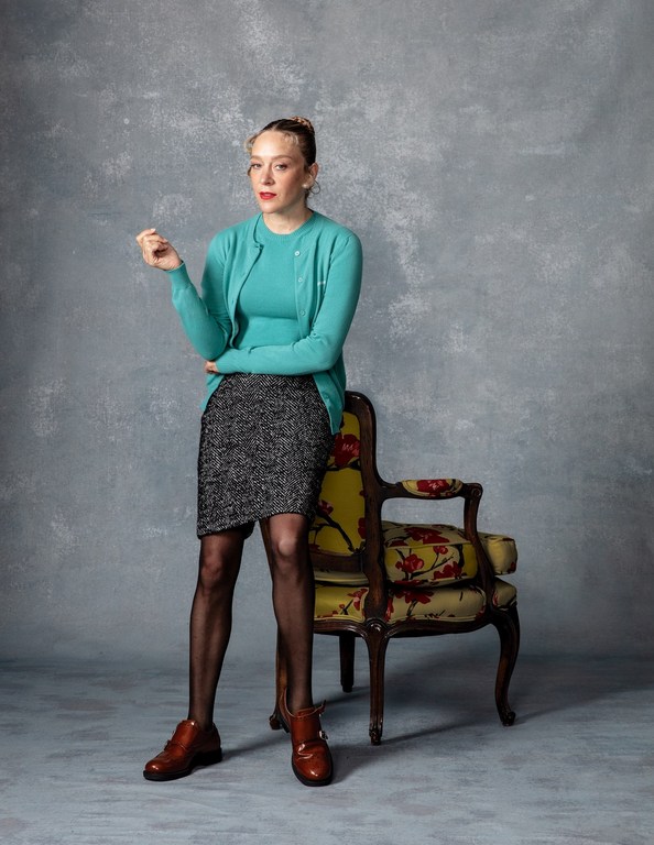A young adult woman in a teal cardigan and pencil skirt stands in front of a mottled grey backdrop, resting her arm on a patterned chair.