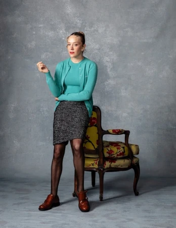 A young adult woman in a teal cardigan and pencil skirt stands in front of a mottled grey backdrop, resting her arm on a patterned chair.