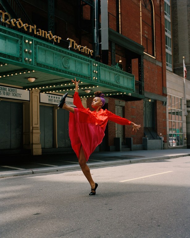 A woman in a red dress strikes a ballet pose in front of the Nederlander Theatre on a city street with brick buildings.