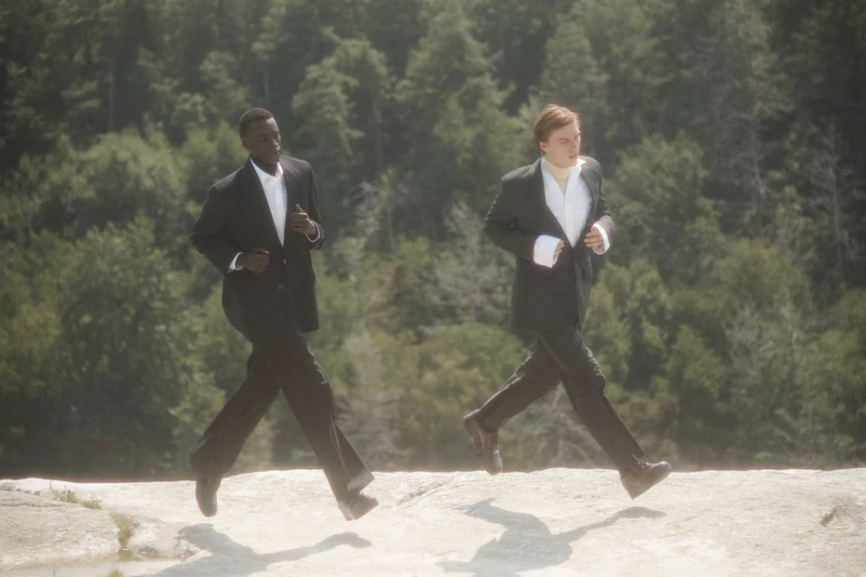 Two men in suits run across a rocky surface with a forest background. The photograph has a soft, dreamy quality and muted colors.