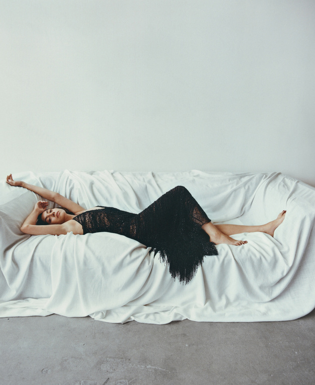 Adult woman in a black lacy dress lies on a white sofa in a minimalist room, showcasing fashion and beauty in a studio setting.