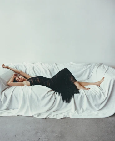 Adult woman in a black lacy dress lies on a white sofa in a minimalist room, showcasing fashion and beauty in a studio setting.