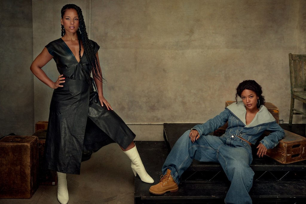Two women pose indoors: one in a leather dress and boots, the other in denim with work boots, sitting on vintage props against a textured wall.