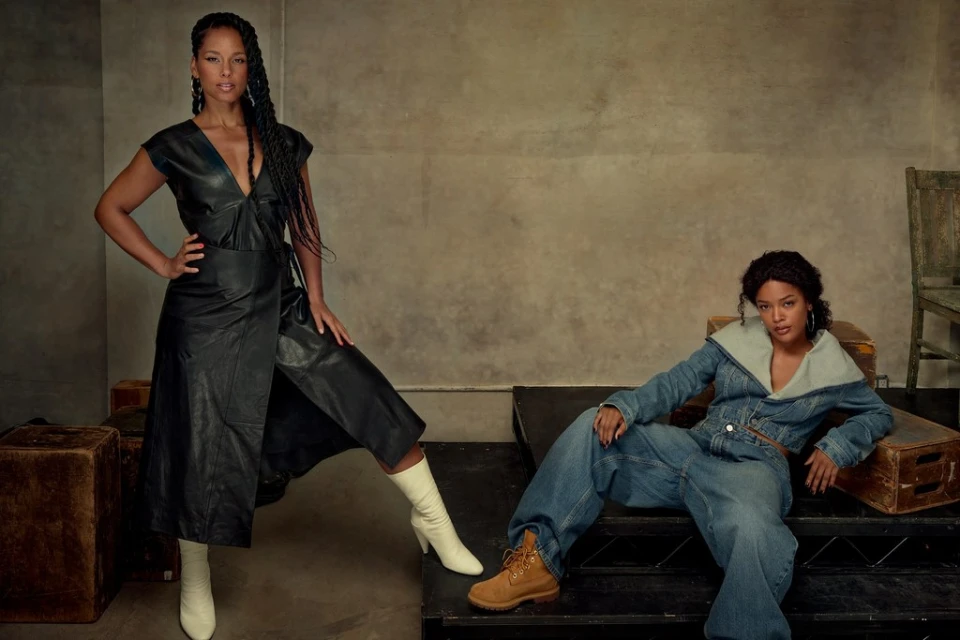Two women pose indoors: one in a leather dress and boots, the other in denim with work boots, sitting on vintage props against a textured wall.