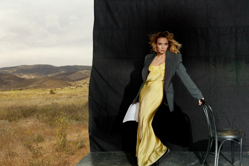 Woman in yellow dress and blazer posing with chair against backdrop of black fabric and outdoor mountain landscape. Studio setting.