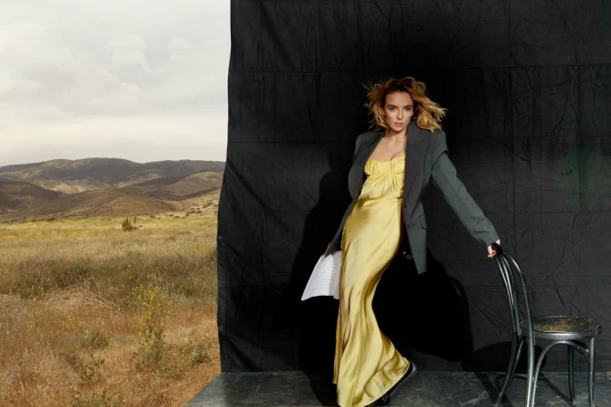 Woman in yellow dress and blazer posing with chair against backdrop of black fabric and outdoor mountain landscape. Studio setting.