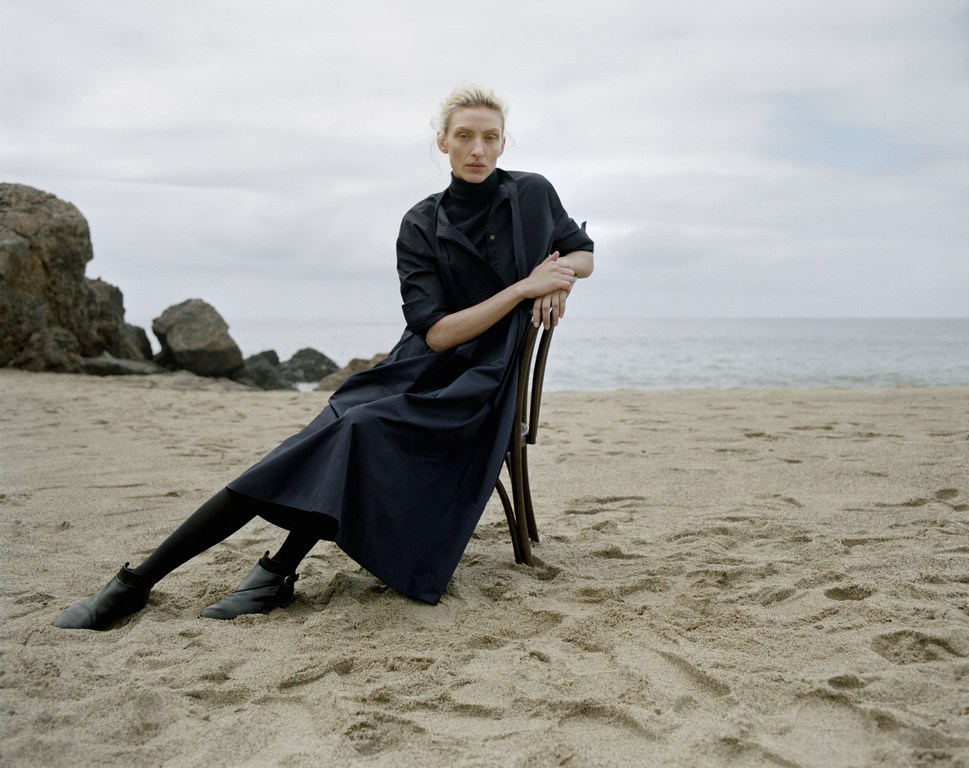 Model wearing dark dress seated on a wooden chair on a sandy beach under an overcast sky.
