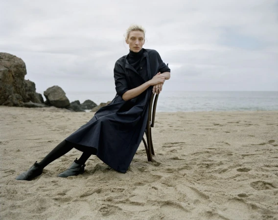 Model wearing dark dress seated on a wooden chair on a sandy beach under an overcast sky.
