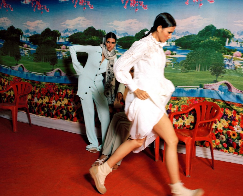 Two female fashion models pose in a red room against a painted landscape backdrop in a fashion editorial shot with unique style.
