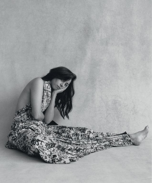 Black and white portrait of a woman in patterned clothing, seated on the floor in a photo studio with her head resting on her hand.