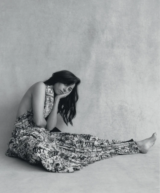 Black and white portrait of a woman in patterned clothing, seated on the floor in a photo studio with her head resting on her hand.