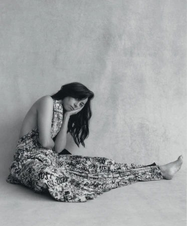 Black and white portrait of a woman in patterned clothing, seated on the floor in a photo studio with her head resting on her hand.