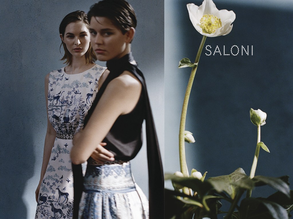 Two women in dresses pose in front of a blue wall next to a white flower with the word "SALONI" printed on the background.