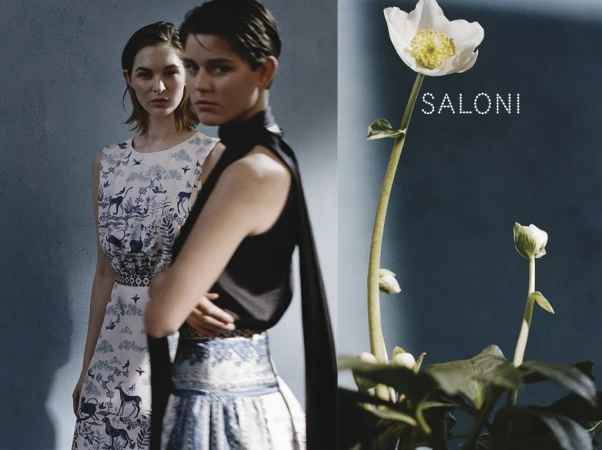 Two women in dresses pose in front of a blue wall next to a white flower with the word "SALONI" printed on the background.