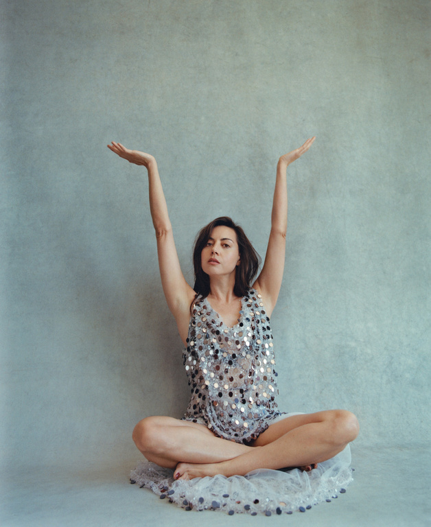 A woman with dark hair sits cross-legged on a sequined surface, arms raised in a studio portrait with a muted blue-gray backdrop.