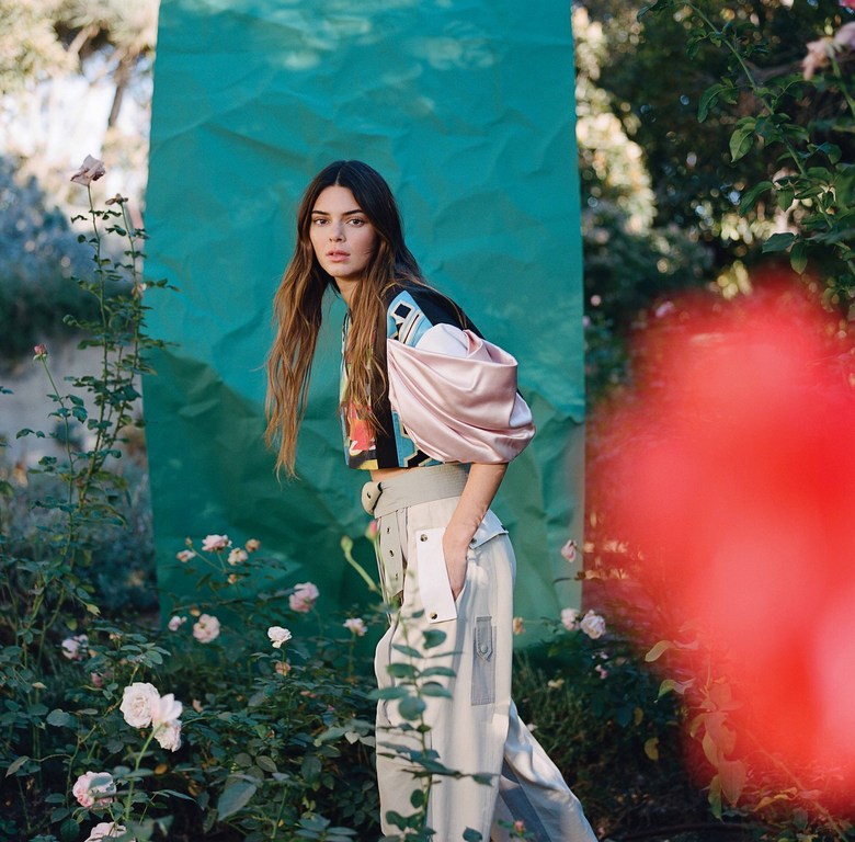 Woman poses in a garden wearing pale trousers, a black-and-pink top, and has long brown hair in an outdoor setting with flowers and greenery.