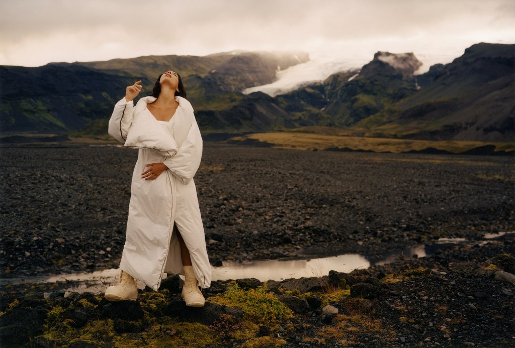 A woman in a white coat and boots poses in a rocky landscape with mountains, and a glacier, set under an overcast sky. Fashion editorial.