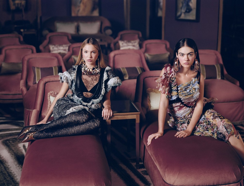 Two female models seated on plush velvet armchairs in a dark theatre wearing ornate patterned dresses and statement jewellery.