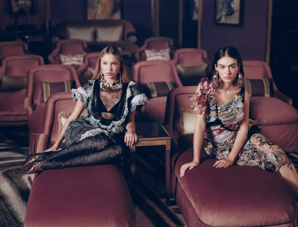 Two female models seated on plush velvet armchairs in a dark theatre wearing ornate patterned dresses and statement jewellery.
