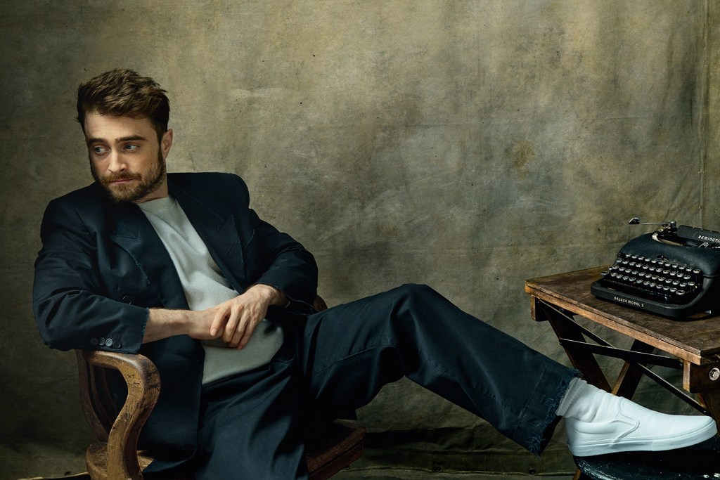 Portrait of a man seated in a wooden chair, wearing a black suit, grey shirt, and white slip-on shoes, with a vintage typewriter nearby.
