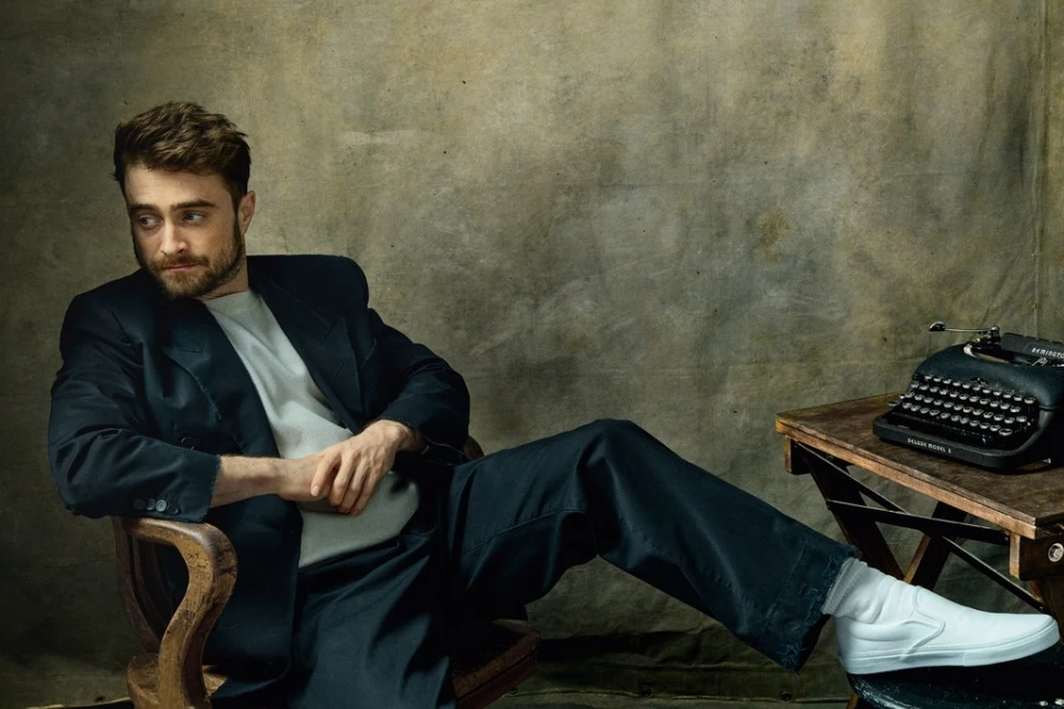 Portrait of a man seated in a wooden chair, wearing a black suit, grey shirt, and white slip-on shoes, with a vintage typewriter nearby.
