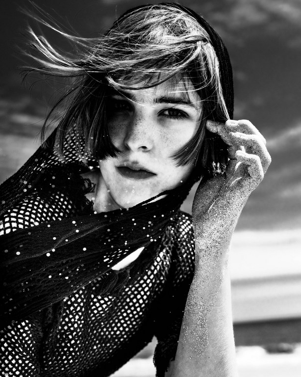 Black and white portrait of a model wearing a fishnet top and hood with sand on her arm and hand against a sky background.