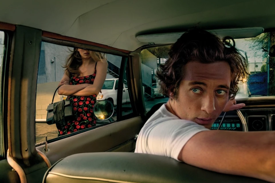 Eye-level shot inside a car with a man in a white t-shirt looking over his shoulder, and a woman in a cherry-print dress visible outside.