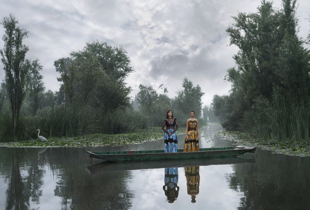 Two women in traditional dresses stand in a wooden boat on a misty river surrounded by lush green trees and vegetation under a cloudy sky.