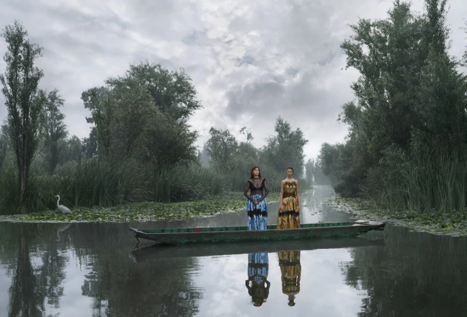 Two women in traditional dresses stand in a wooden boat on a misty river surrounded by lush green trees and vegetation under a cloudy sky.