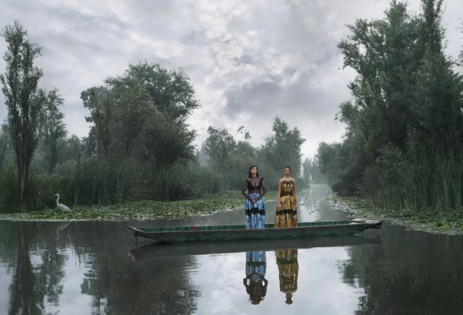 Two women in traditional dresses stand in a wooden boat on a misty river surrounded by lush green trees and vegetation under a cloudy sky.