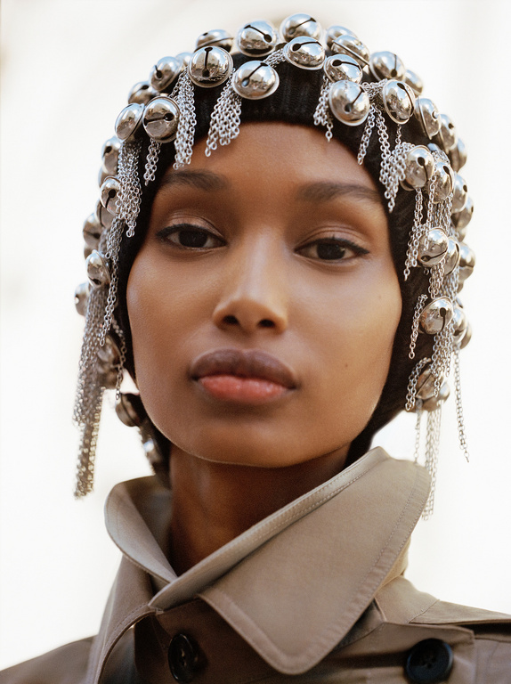 Close-up of a model with a tan trench coat and a silver jingle bell hat against an off-white background in a fashion style shot.