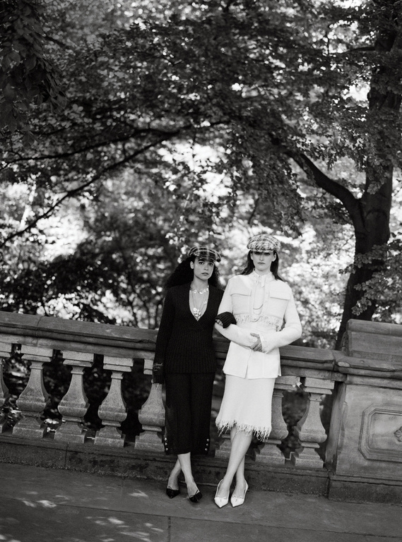 Black and white photograph of two women in stylish suits and hats posing in front of a stone railing with trees in the background.