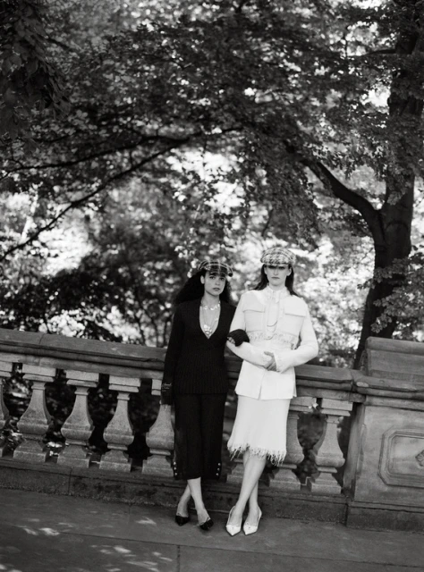 Black and white photograph of two women in stylish suits and hats posing in front of a stone railing with trees in the background.