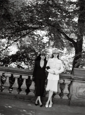 Black and white photograph of two women in stylish suits and hats posing in front of a stone railing with trees in the background.