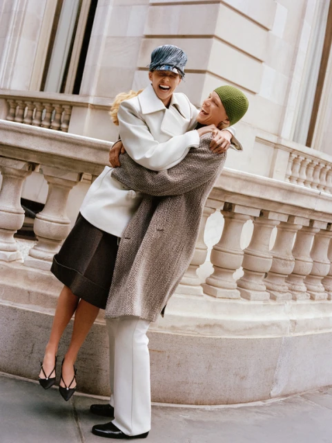 Happy couple embracing outdoors near stone architecture. Woman in hat and coat, being lifted by man in white pants and coat.