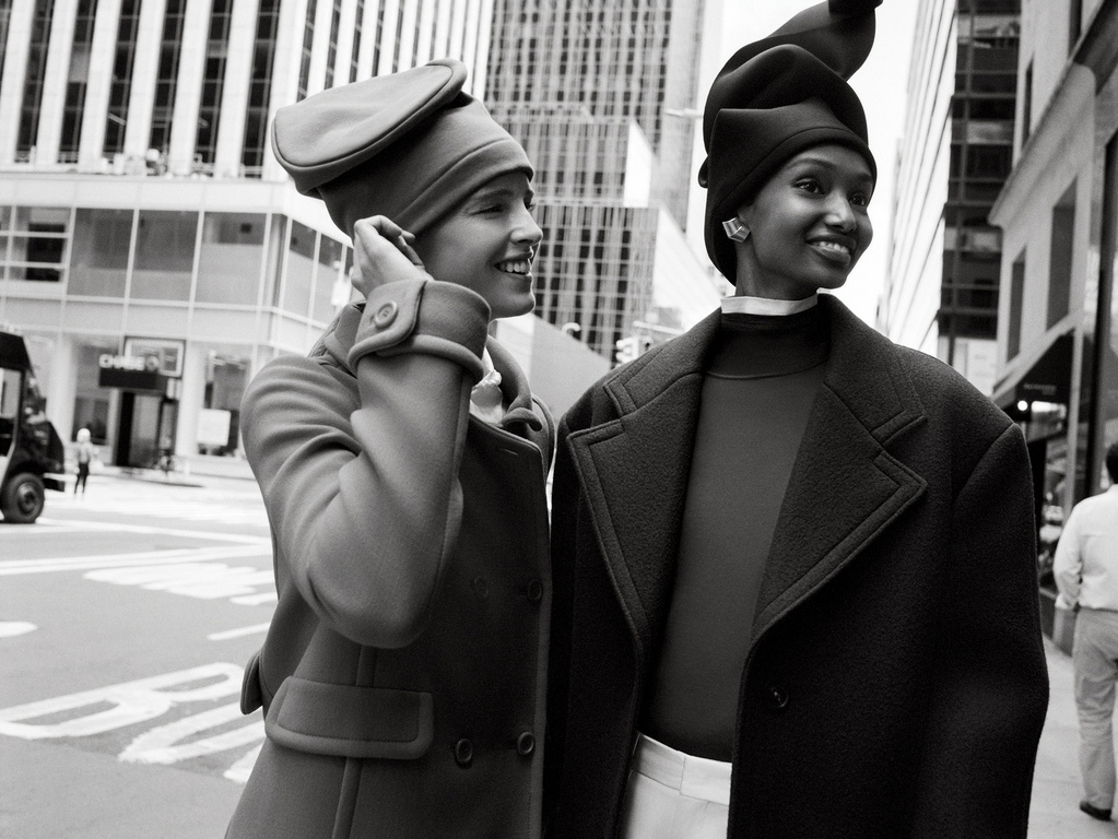 Two stylish women model vintage-inspired coats and hats in a monochrome outdoor fashion shot on a city street with skyscrapers.