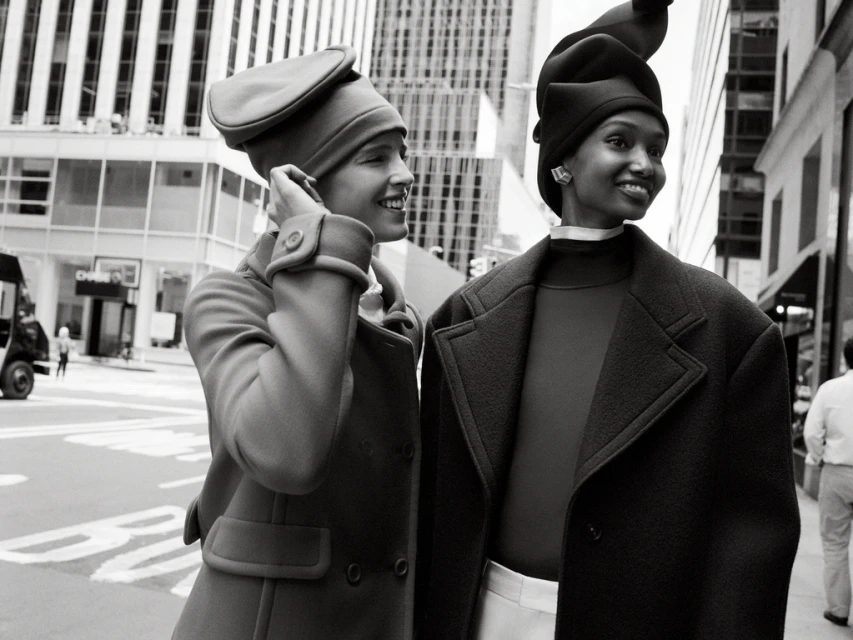 Two stylish women model vintage-inspired coats and hats in a monochrome outdoor fashion shot on a city street with skyscrapers.