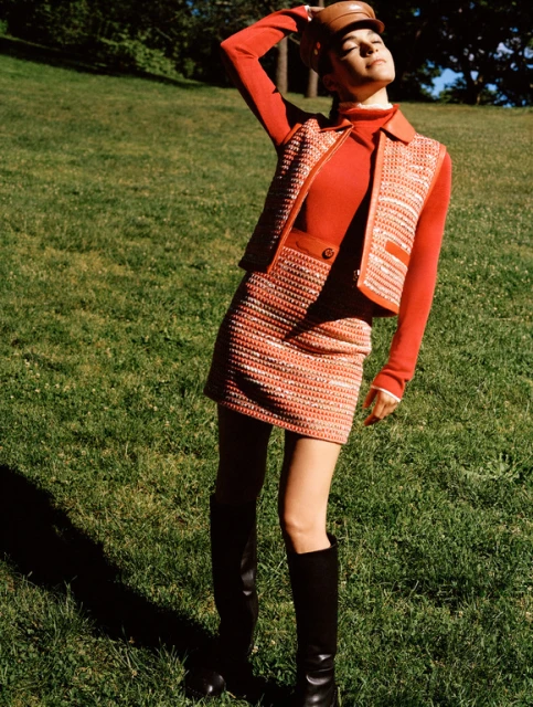 A woman poses in a retro-style orange tweed skirt and vest with black boots on a grassy hill in a natural, outdoor setting.
