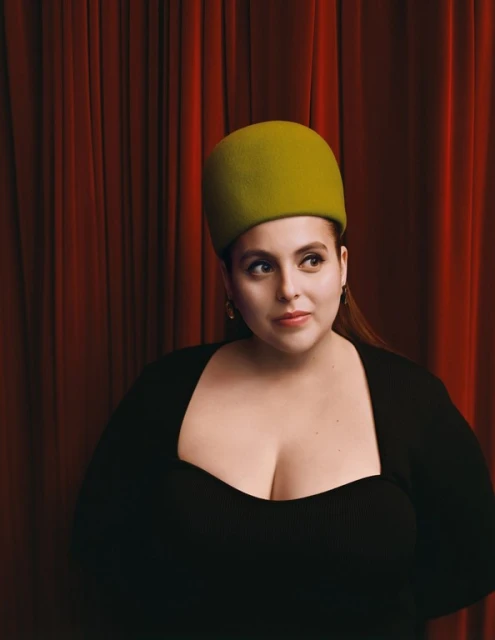Portrait of an adult female with long brown hair wearing a green hat and black top, posing in front of a red curtain background.