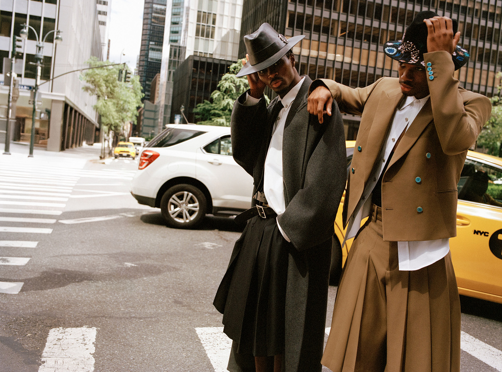 Two stylish men in hats and skirts pose on a city street with a white SUV and a yellow taxi cab in the background in an urban setting.