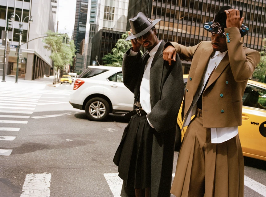 Two stylish men in hats and skirts pose on a city street with a white SUV and a yellow taxi cab in the background in an urban setting.