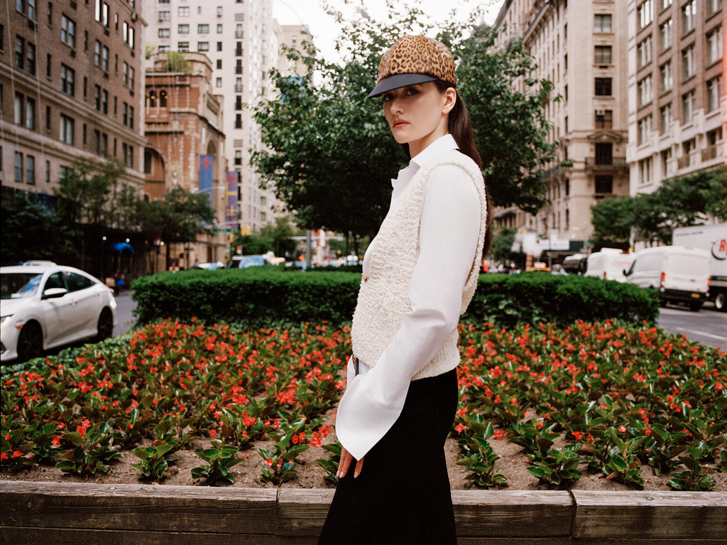 A woman in a printed cap and white vest poses on a city street lined with buildings, cars, trees, and a flower bed in an editorial style photograph.