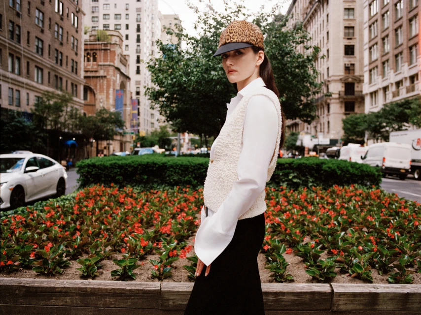 A woman in a printed cap and white vest poses on a city street lined with buildings, cars, trees, and a flower bed in an editorial style photograph.