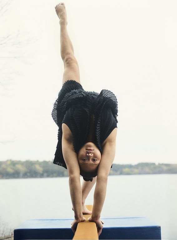 Young woman in a black dress balances on a beam, one leg raised, outdoors with a lake and trees in the background, capturing a moment of strength.