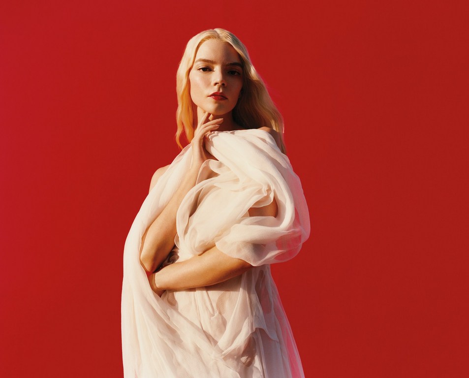 Anya Taylor-Joy poses in a flowing white gown against a vibrant red backdrop, illuminated by soft, directional lighting for a fashion editorial shot.